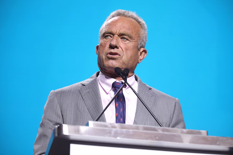 A confident man in a suit delivers a speech at a podium, with a bright blue background.