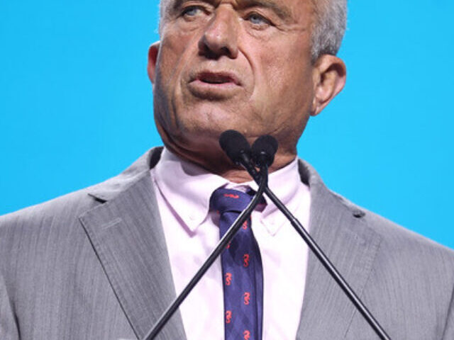 A confident man in a suit delivers a speech at a podium, with a bright blue background.