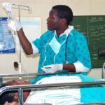 A doctor, attending a patient in a hospital setting. The healthcare worker is dressed in a teal-colored medical uniform with gloves and a stethoscope, adjusting an IV drip for a patient lying on a hospital bed. Photo Source: JacobOcenFay (CC BY-SA 4.0)