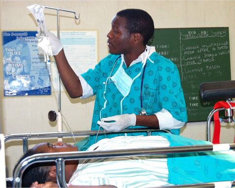 A doctor, attending a patient in a hospital setting. The healthcare worker is dressed in a teal-colored medical uniform with gloves and a stethoscope, adjusting an IV drip for a patient lying on a hospital bed. Photo Source: JacobOcenFay (CC BY-SA 4.0)