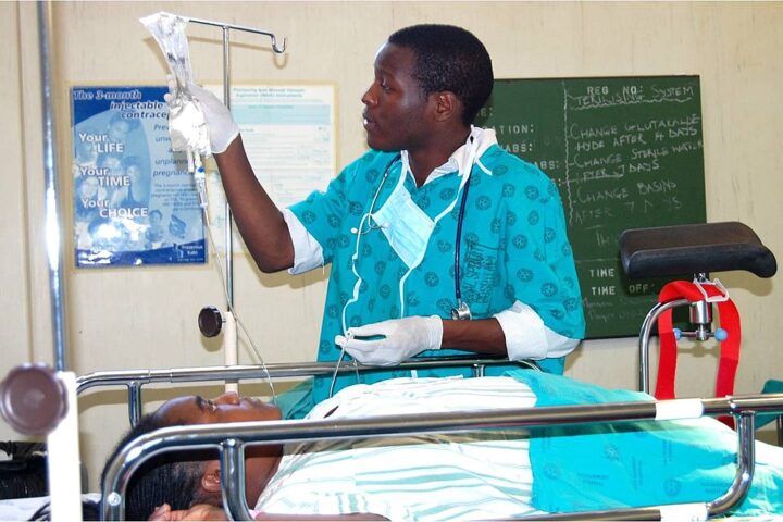 A doctor, attending a patient in a hospital setting. The healthcare worker is dressed in a teal-colored medical uniform with gloves and a stethoscope, adjusting an IV drip for a patient lying on a hospital bed. Photo Source: JacobOcenFay (CC BY-SA 4.0)