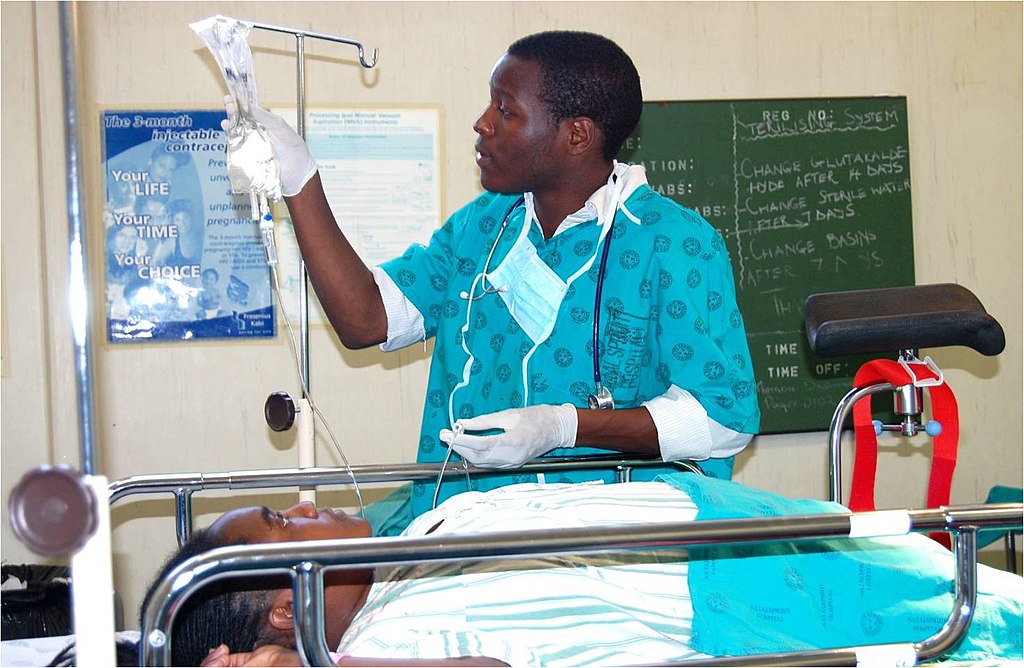 A doctor, attending a patient in a hospital setting. The healthcare worker is dressed in a teal-colored medical uniform with gloves and a stethoscope, adjusting an IV drip for a patient lying on a hospital bed. Photo Source: JacobOcenFay (CC BY-SA 4.0)