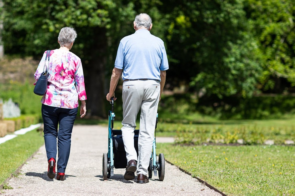 Representative Image: Two People Walking in a Park. Elderly couple walking along a pathway in a park or garden. The man is using a walker for support, while the woman walks beside him. Photo Source: Rollz International (Pexels)