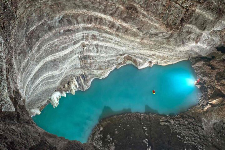 Two kayakers explore a glowing blue pool in a vast, rocky cave. Photo Source: Nadace Neuron