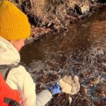 Person in yellow beanie examines foam near partially frozen waterway. Photo Source: UNH Research Team.