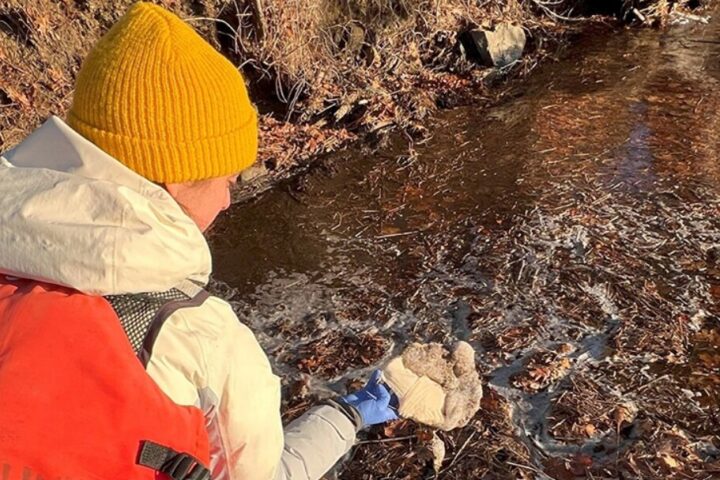 Person in yellow beanie examines foam near partially frozen waterway. Photo Source: UNH Research Team.