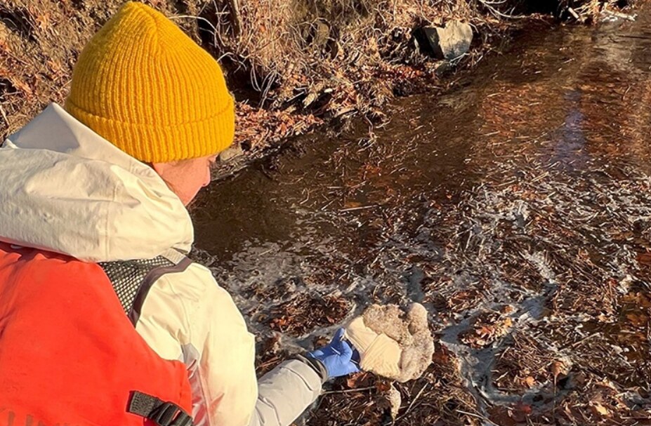 Person in yellow beanie examines foam near partially frozen waterway. Photo Source: UNH Research Team.