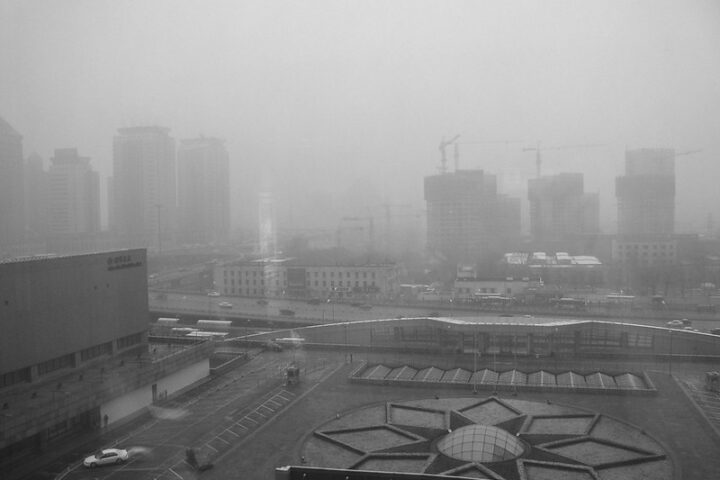 Representative Image: A cityscape covered in heavy smog, with high-rise buildings and construction cranes barely visible in the background. The scene suggests severe air pollution, likely due to industrial activities or coal dependency. Photo Source: Kevin Dooley (CC BY 2.0)