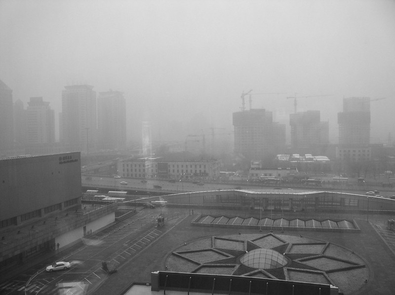 Representative Image: A cityscape covered in heavy smog, with high-rise buildings and construction cranes barely visible in the background. The scene suggests severe air pollution, likely due to industrial activities or coal dependency. Photo Source: Kevin Dooley (CC BY 2.0)