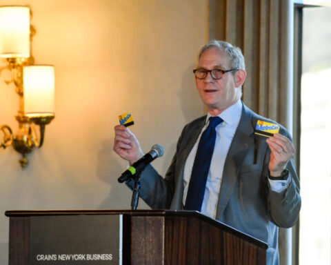 A person standing at a podium, speaking into a microphone, and holding up two MetroCards. Photo Source - MTA