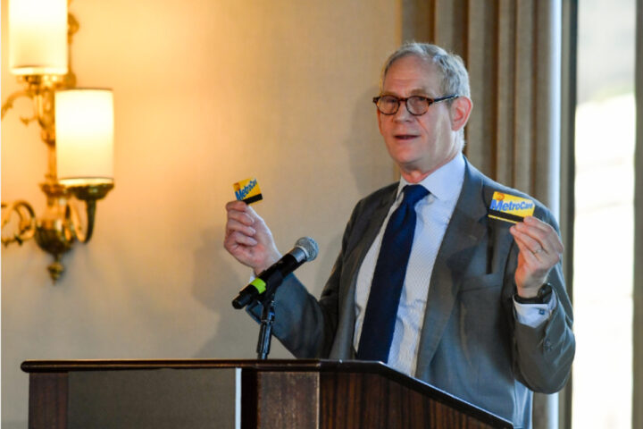 A person standing at a podium, speaking into a microphone, and holding up two MetroCards. Photo Source - MTA