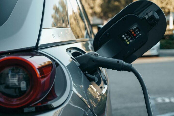 Representative Image: Electric vehicle (EV) being charged. The charging cable is plugged into the car’s charging port, with the port cover open, displaying indicator lights and symbols. Photo Source: Pexel (Kindel Media)