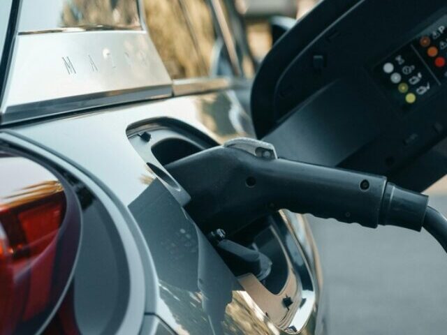 Representative Image: Electric vehicle (EV) being charged. The charging cable is plugged into the car’s charging port, with the port cover open, displaying indicator lights and symbols. Photo Source: Pexel (Kindel Media)